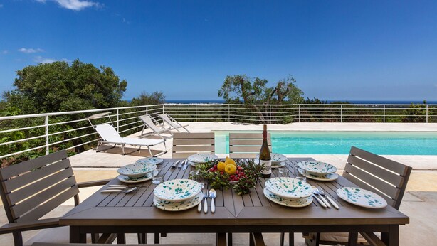 A dining table set for six with plates, cutlery, and a bottle of wine, overlooking a pool and ocean view.