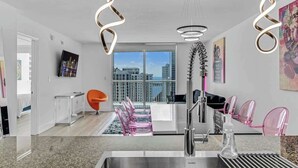 A modern kitchen with a stainless steel sink, a dining area with pink chairs, and a large window offering a city view. - Curated Sky Abode: Ocean & City views, Sleeps 6 (Miami)