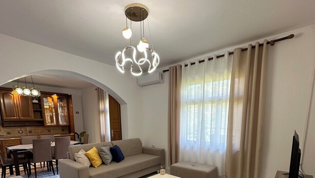 A modern living room with a sofa, coffee table, and TV stand. There is a kitchen area with wooden cabinets and a dining table. - Family - sized villa , pet friendly , city and castle view with a big garden ! (Berat)