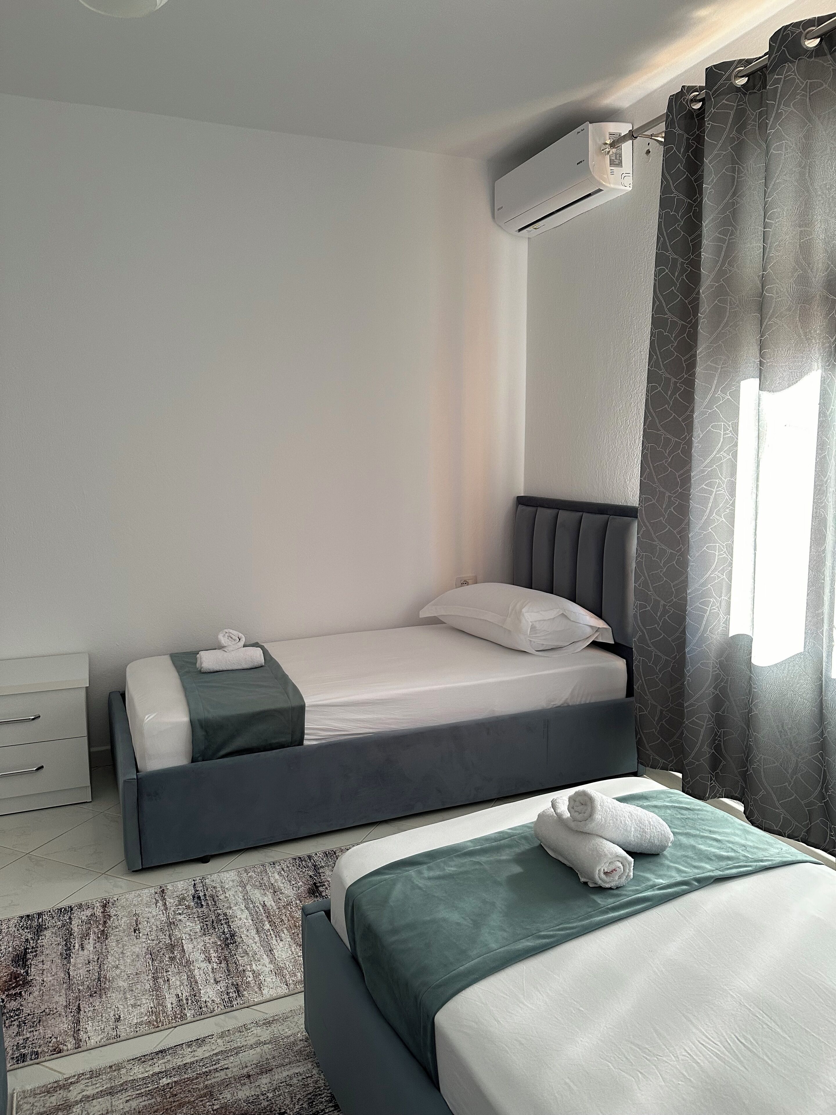 Una camera d'albergo moderna con un letto singolo, un comodino, un condizionatore d'aria e una finestra con tende.