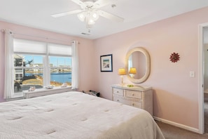 Quarto com cama, ventilador de teto, cômoda, espelho e janela com vista para um corpo de água.