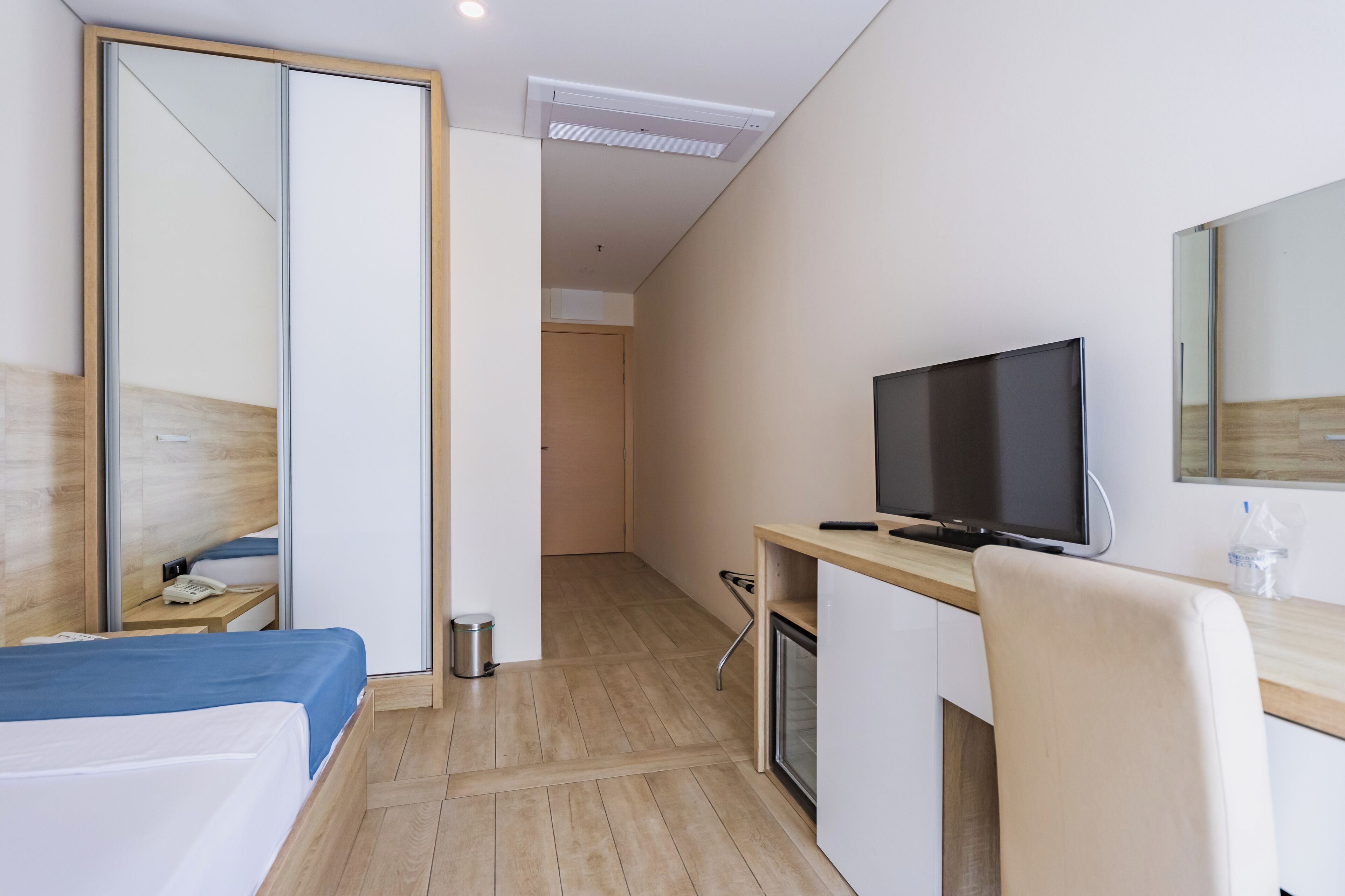 A hotel room with a bed, a desk with a television, a mirror, and a small trash bin.