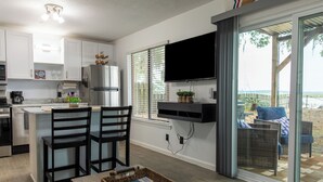 A modern kitchen with white cabinets, a stainless steel refrigerator, and a built-in TV. There is a dining area with two chairs and a table, and a wicker chair outside on a deck. - Romantic Waterfront Getaway
 1st Floor Deep Water Views (Beaufort)