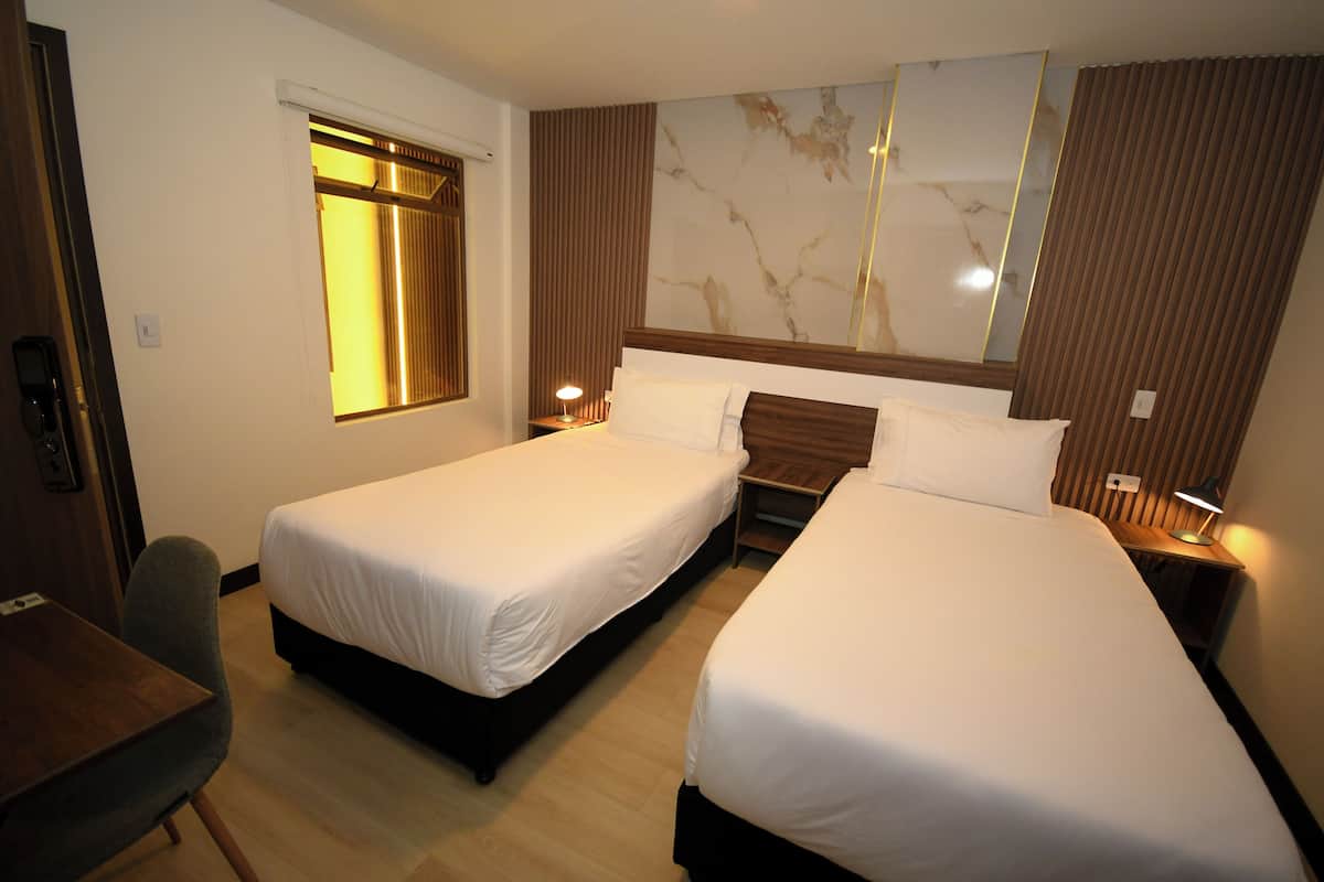 A hotel room with two single beds, a desk, a chair, a window with blinds, and a large mirror on the wall.