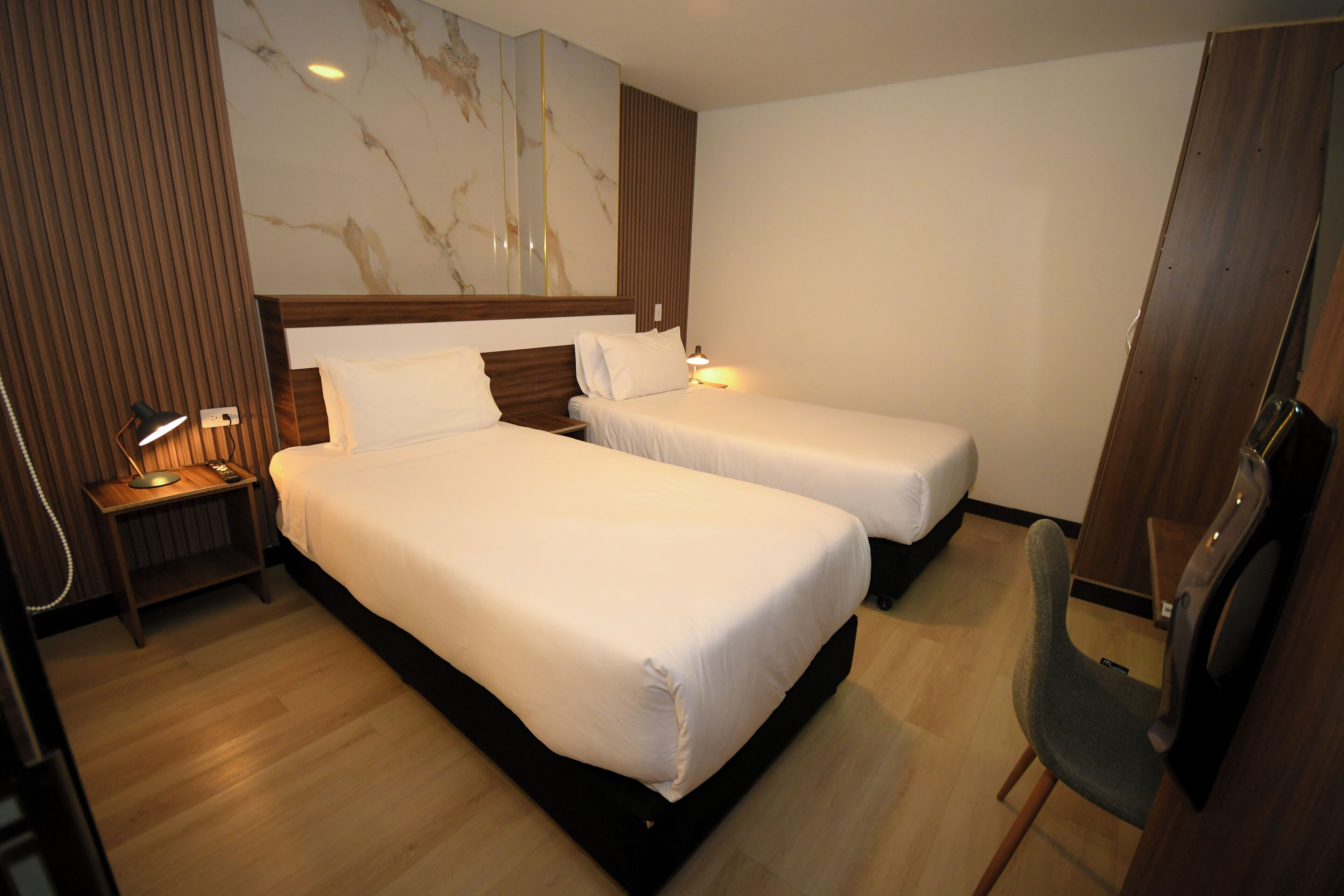 A hotel room with two beds, a wooden nightstand with a lamp, a chair, and a large mirror on the wall.