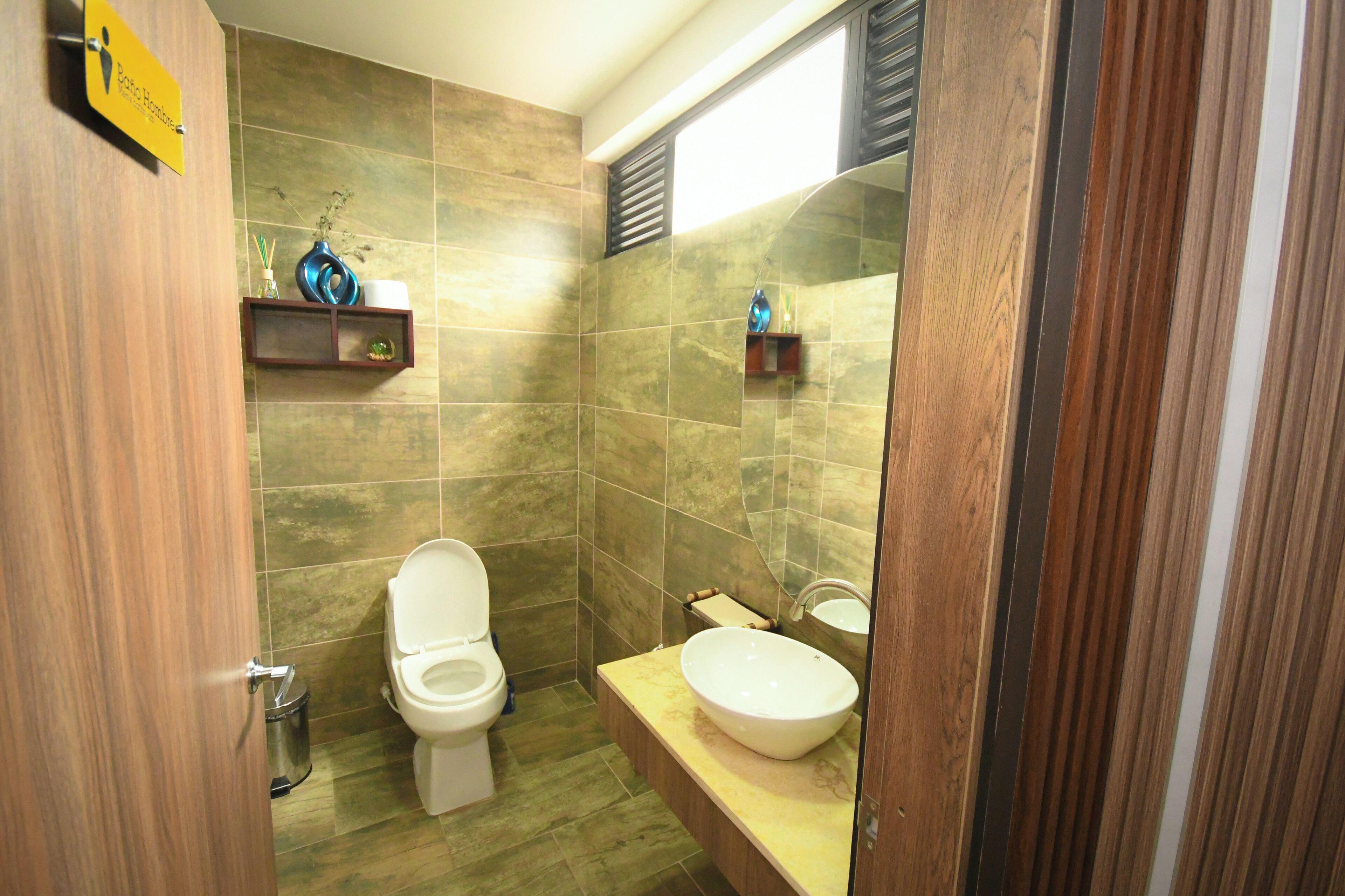 A bathroom with a toilet, sink, and shower area. There are wooden shelves with toiletries.