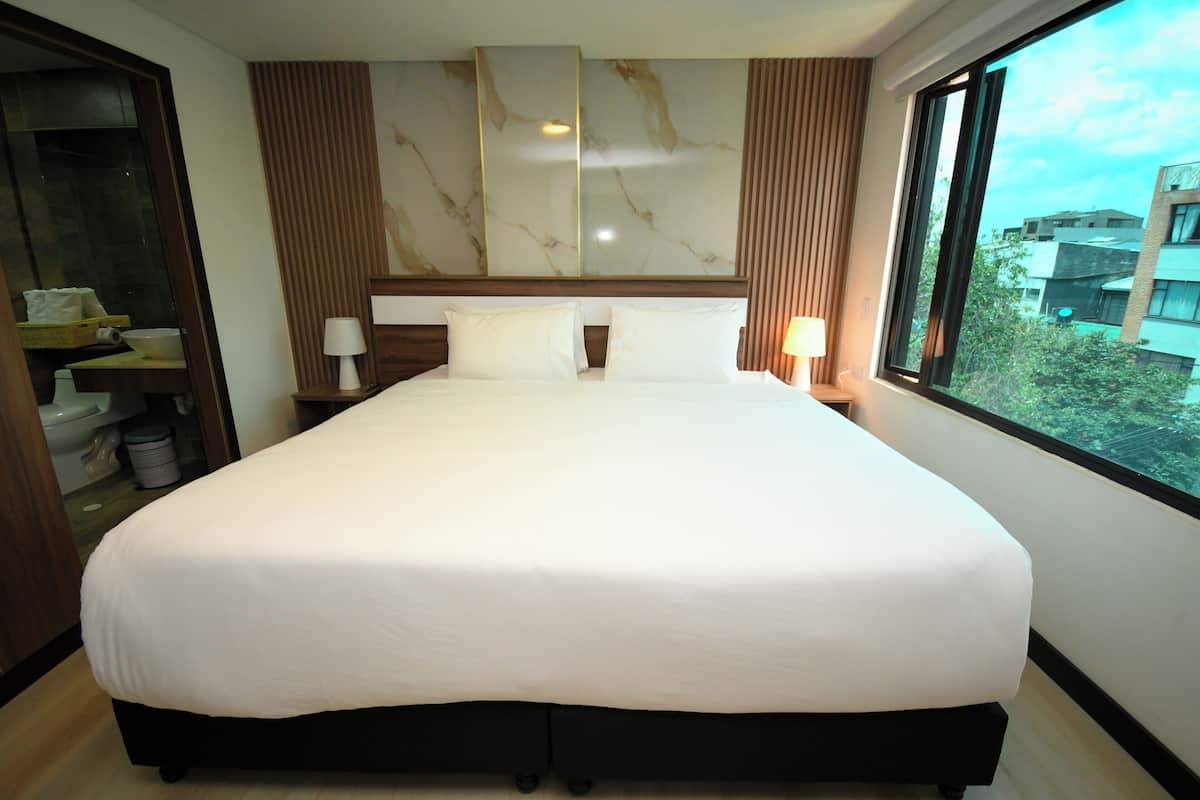 A large bed with white linens, a marble headboard, and a view of buildings through a window.