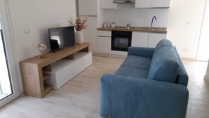 Moderni olohuone, jossa on sininen sohva, puinen TV-taso sekä keittiöalue, jossa on valkoiset kaapit ja liesi.
