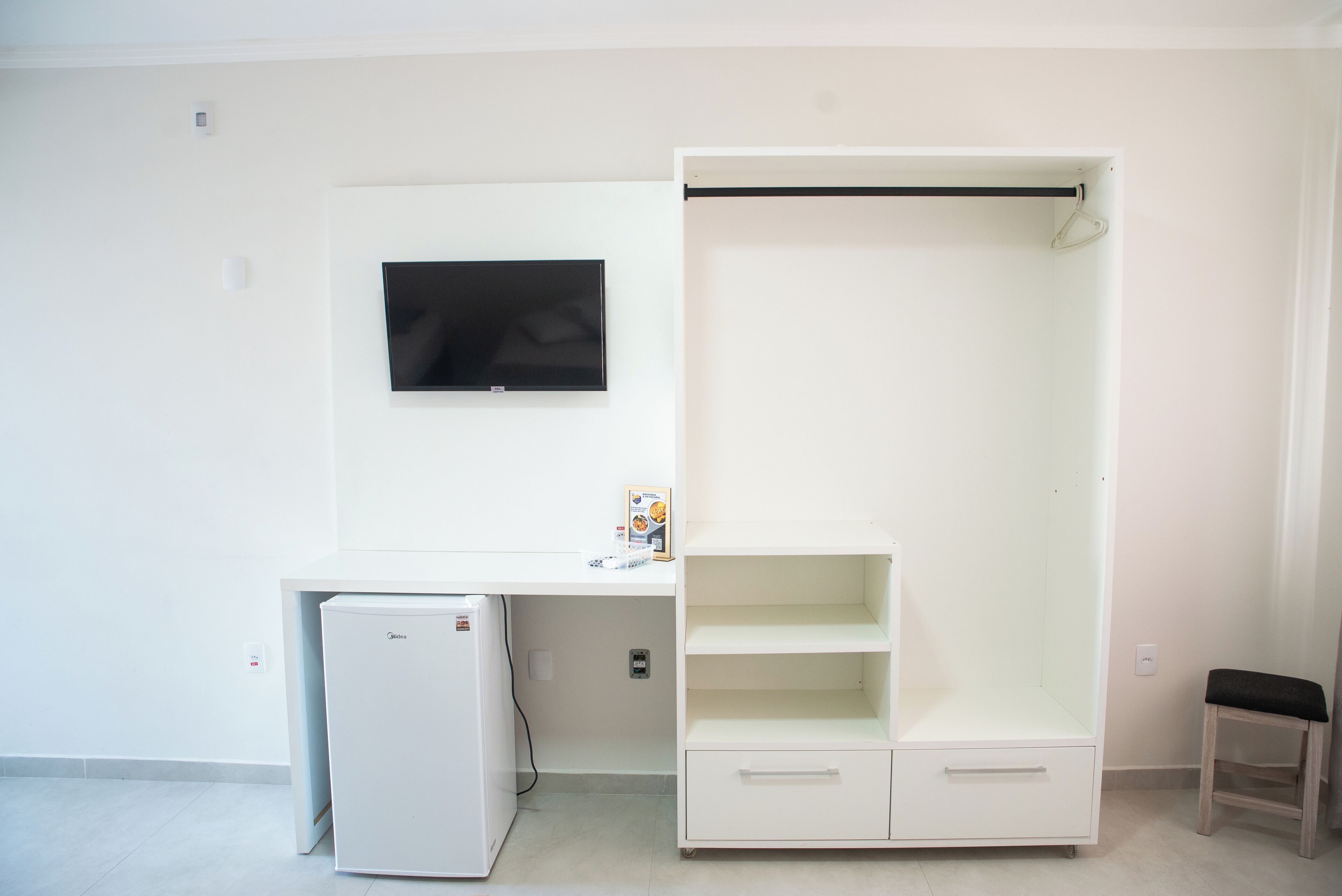 A room with a white desk, a flat-screen TV mounted on the wall, a white wardrobe with a hanging rod, and a small white refrigerator.