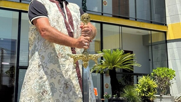A person in a patterned robe holding a sword, standing in front of a building with balconies and potted plants. - POUSADA CASTELO DO REI (Penha)