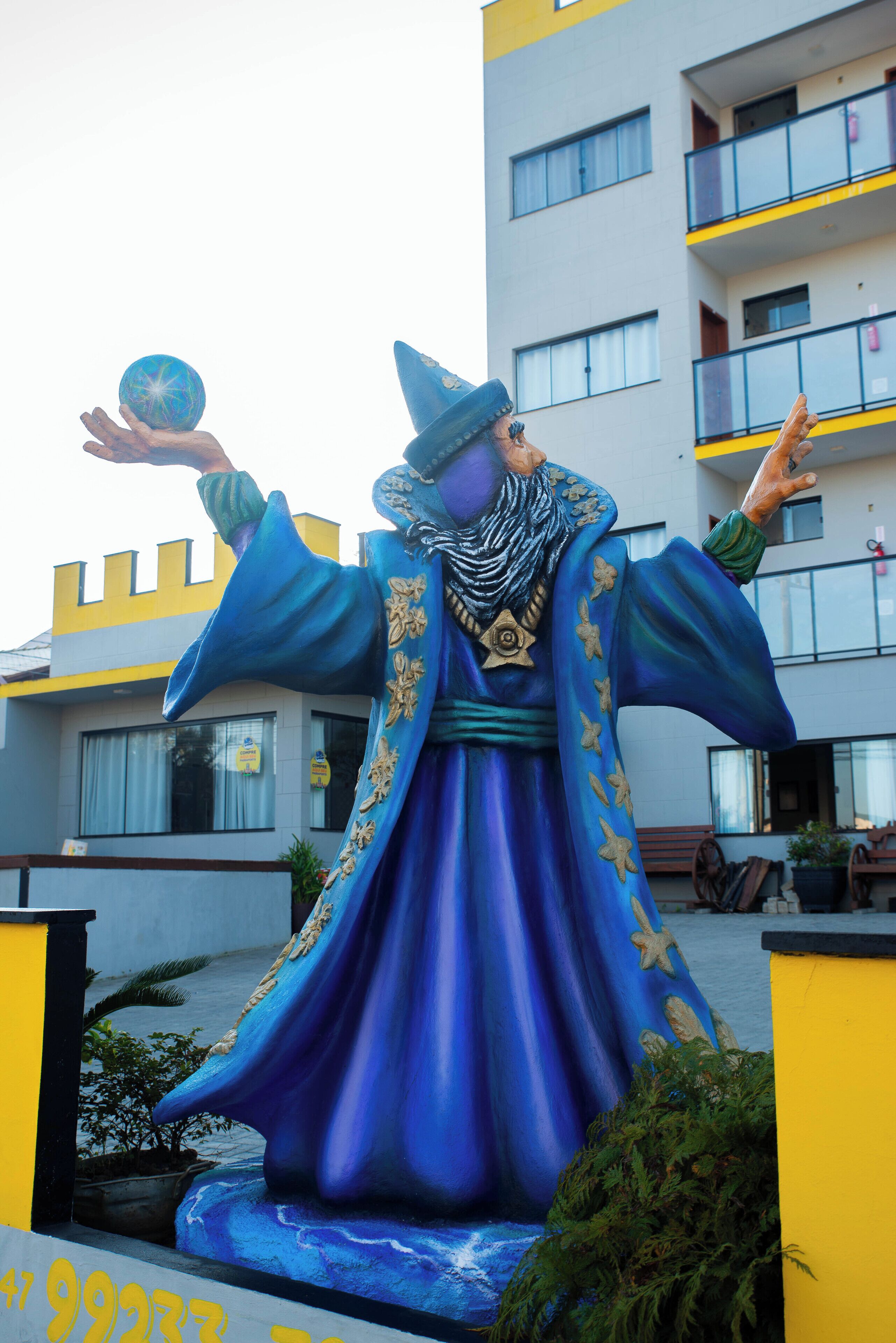 Una estatua de una figura con túnica azul adornada con motivos de estrellas de mar, sosteniendo una esfera de cristal, frente a un edificio con balcones y ventanas.