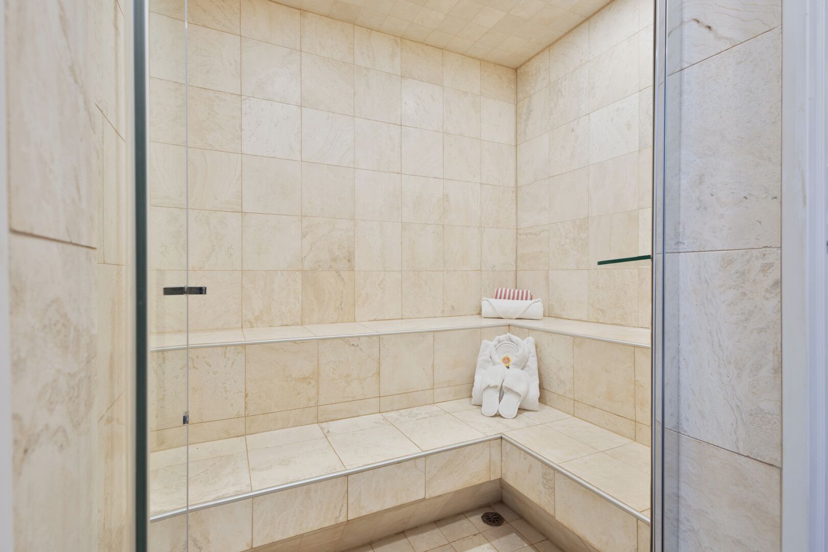 A corner shower with beige tiles, a glass door, and a bench with towels.