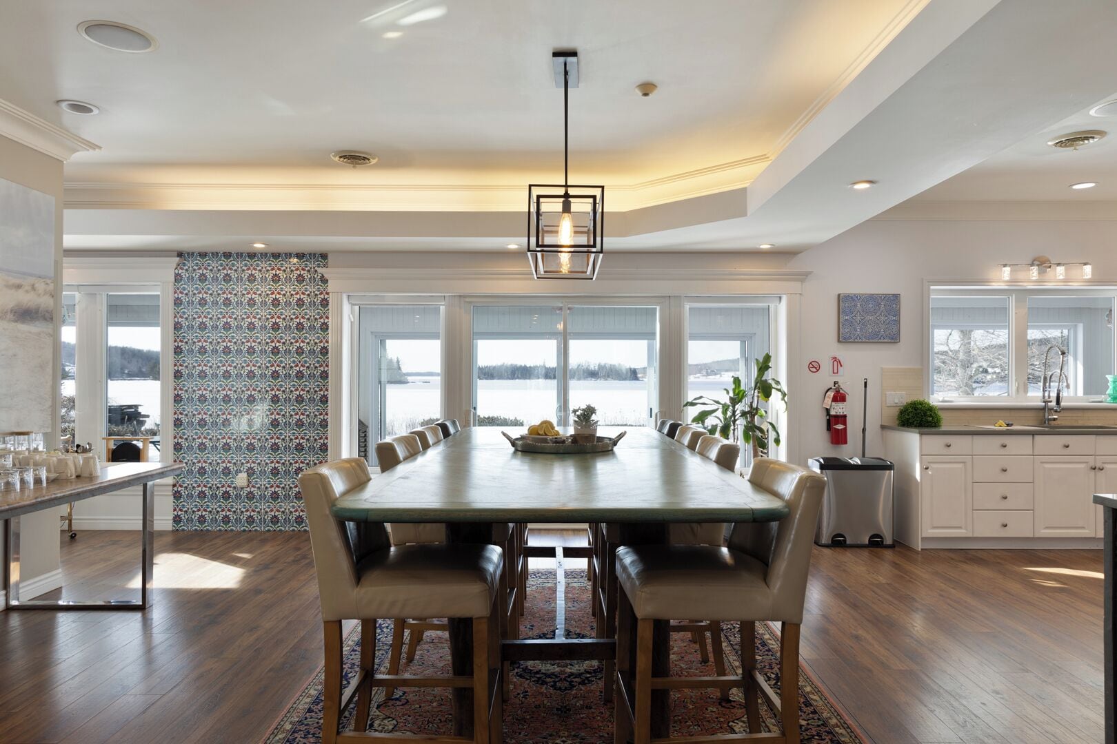 A spacious dining area with a long table, multiple chairs, and a view of a lake through large windows.