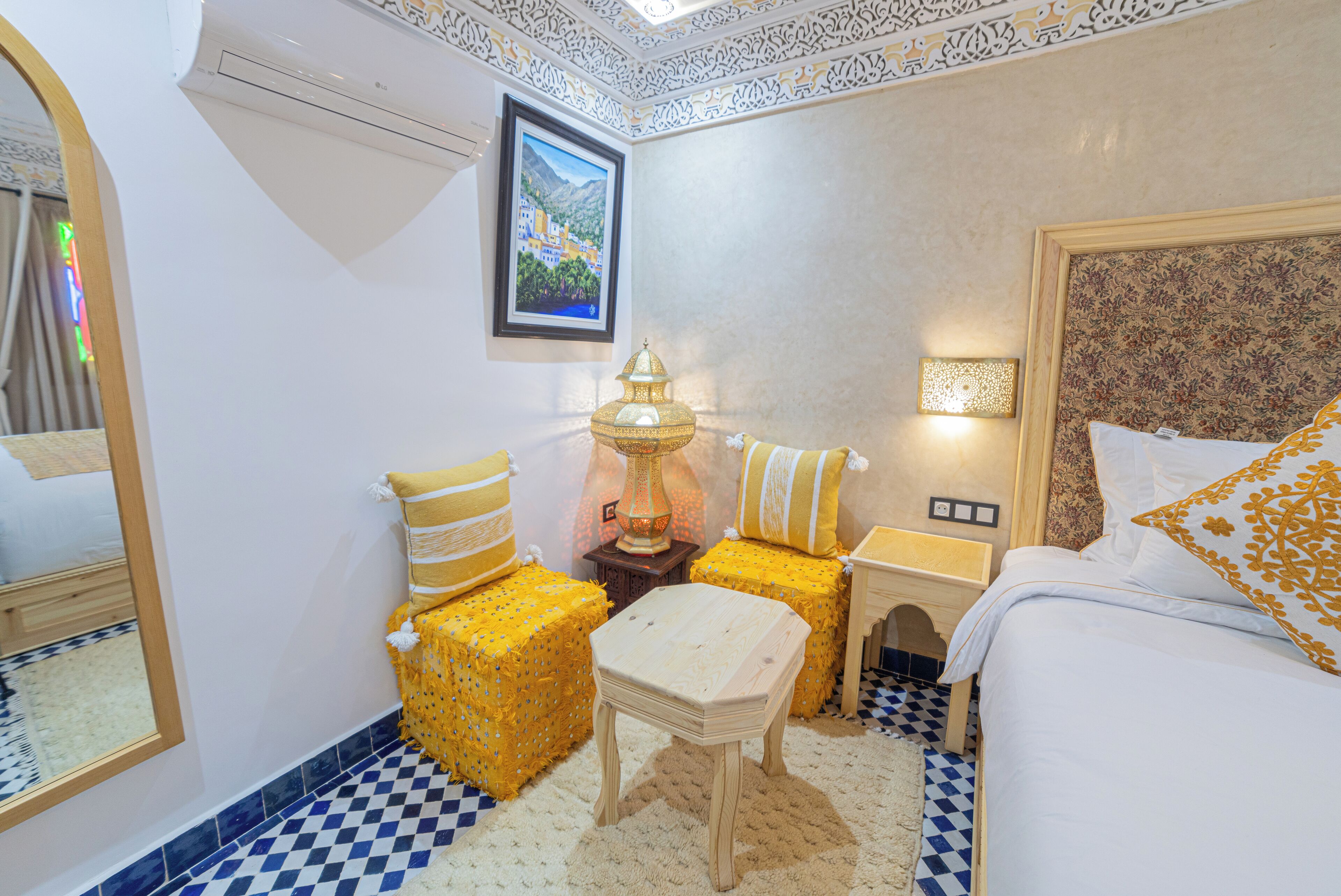A hotel room with a checkered floor, a bed with white and gold pillows, a small wooden table, two yellow cushioned stools, a wall-mounted lamp, and a framed picture above the bed.