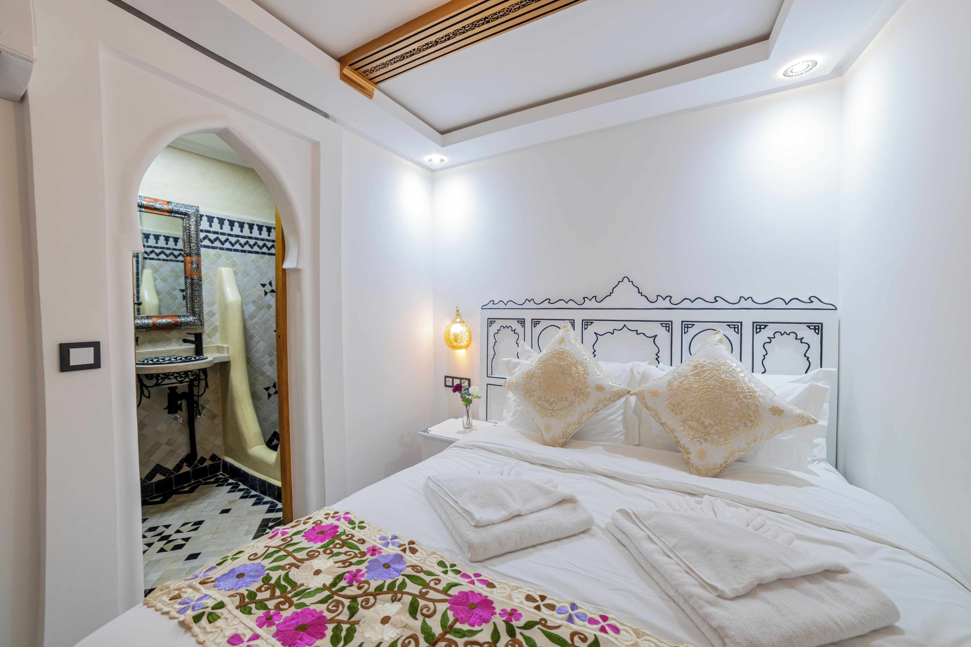 A hotel room with a bed, a nightstand, a wall-mounted lamp, and a bathroom visible through an archway.