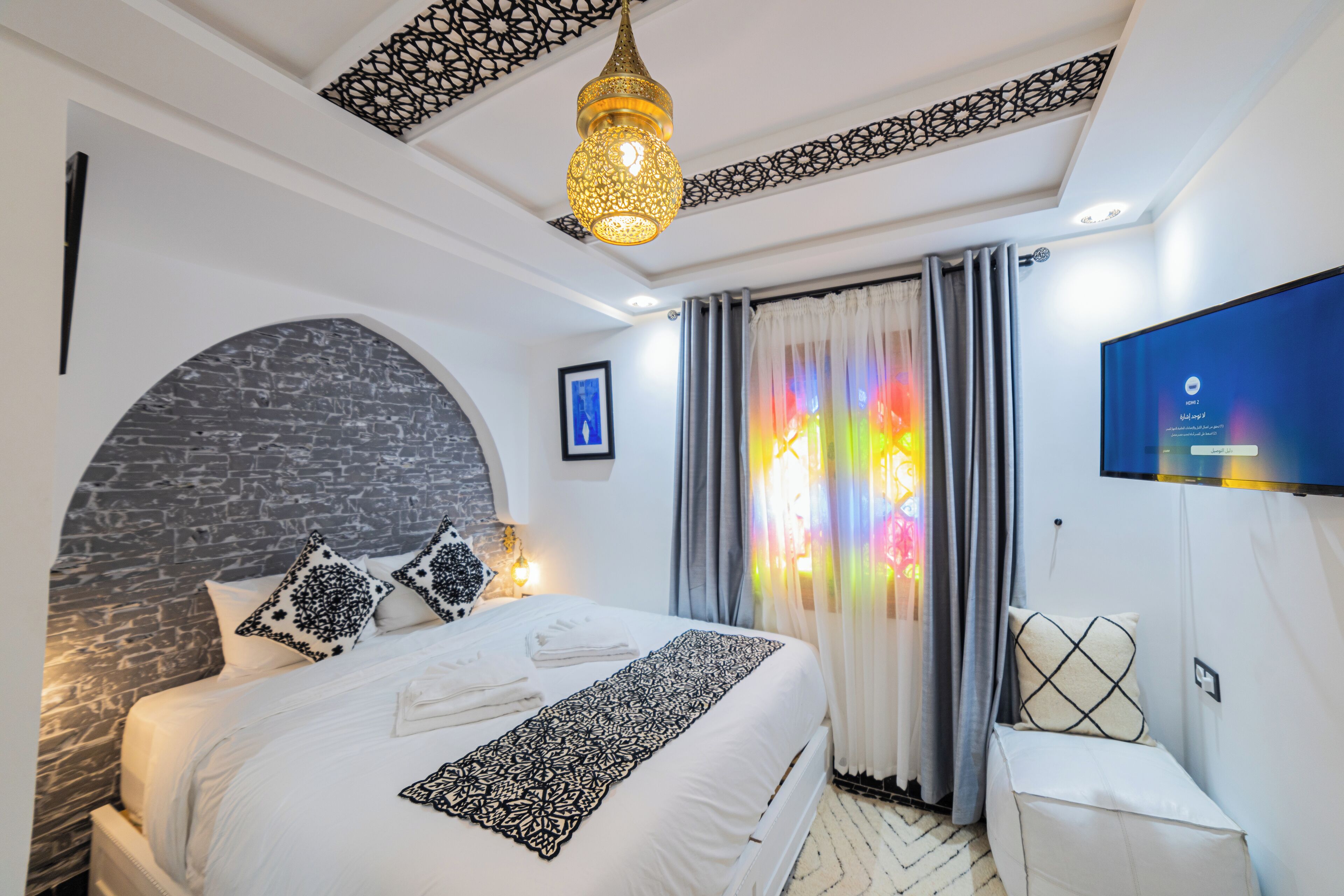 Quarto moderno com cama grande, luminária decorativa no teto, televisão e vista para o exterior através de uma janela com cortinas.