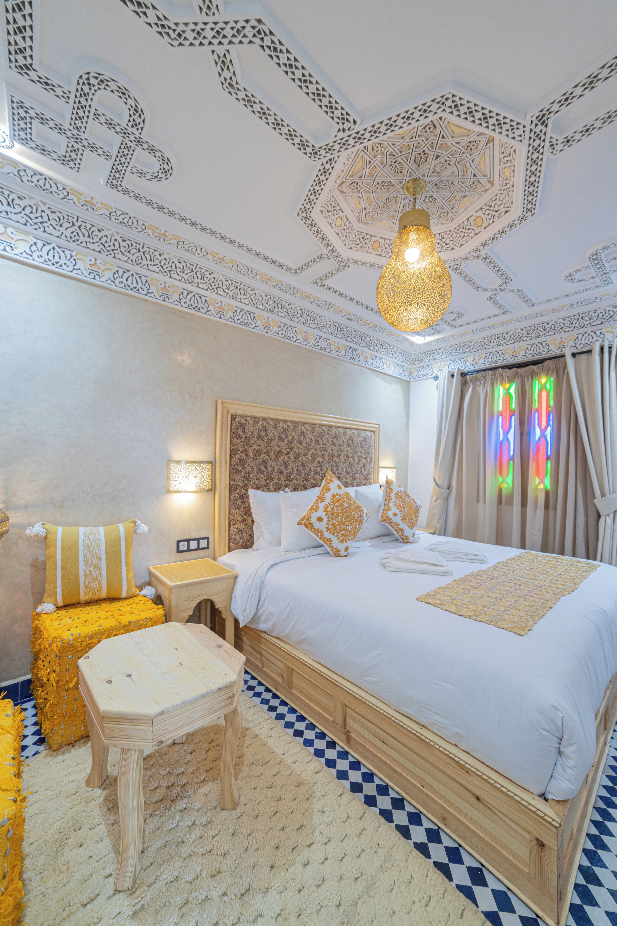 A bedroom with a large bed, a wooden nightstand, a small stool, and a decorative wall light.