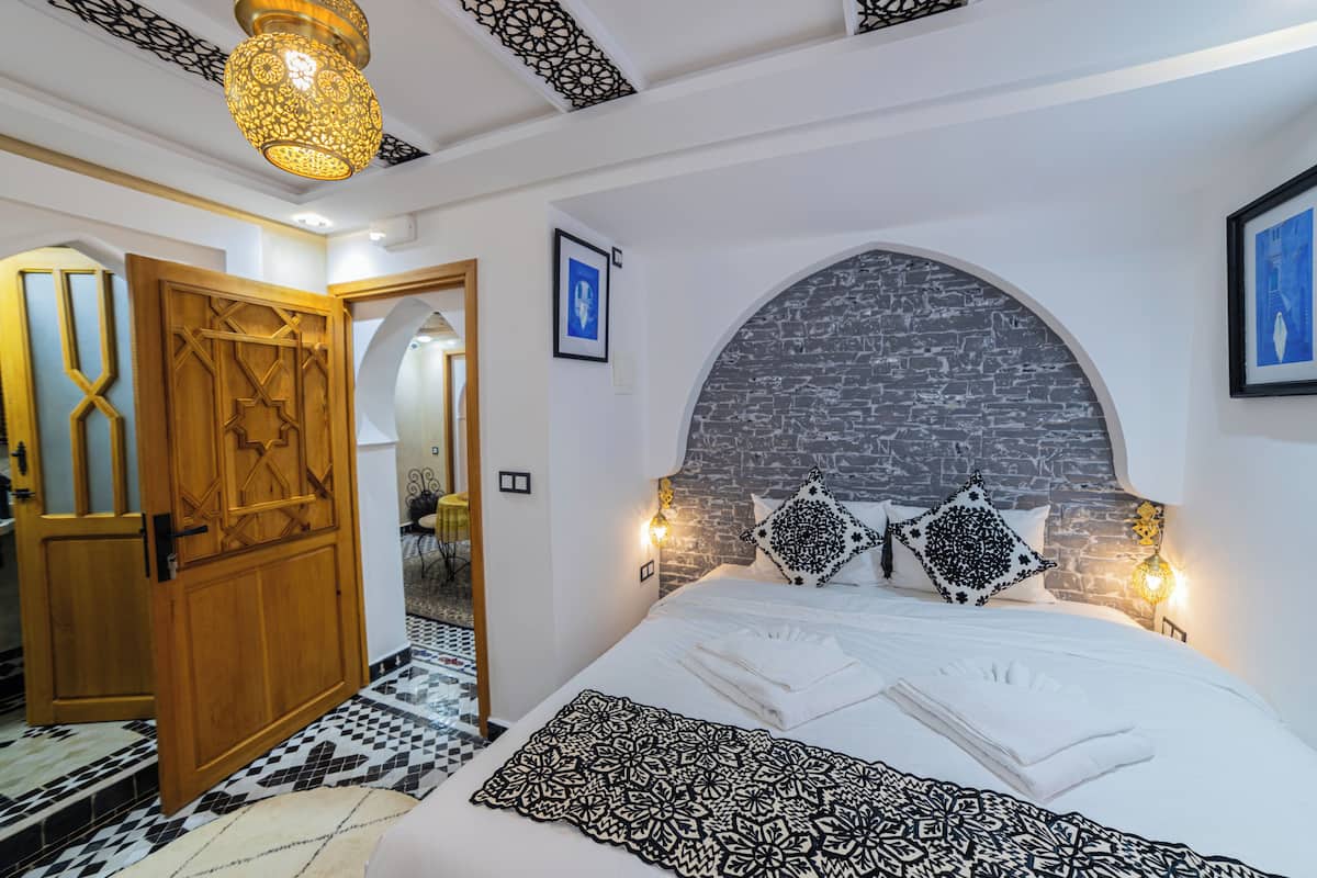 A bedroom with a large bed, a wooden door, a patterned rug, and a stone wall.