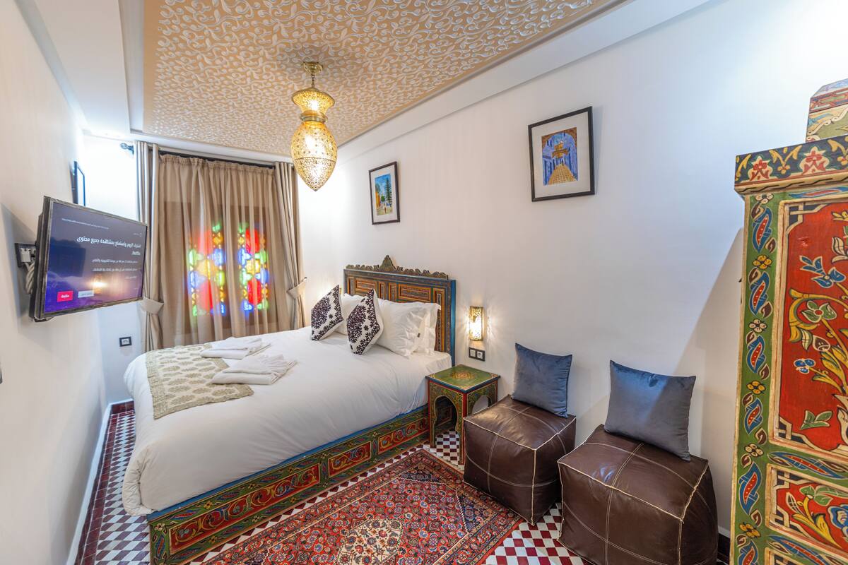A bedroom with a bed, a television, a nightstand, a bench, and a patterned rug.