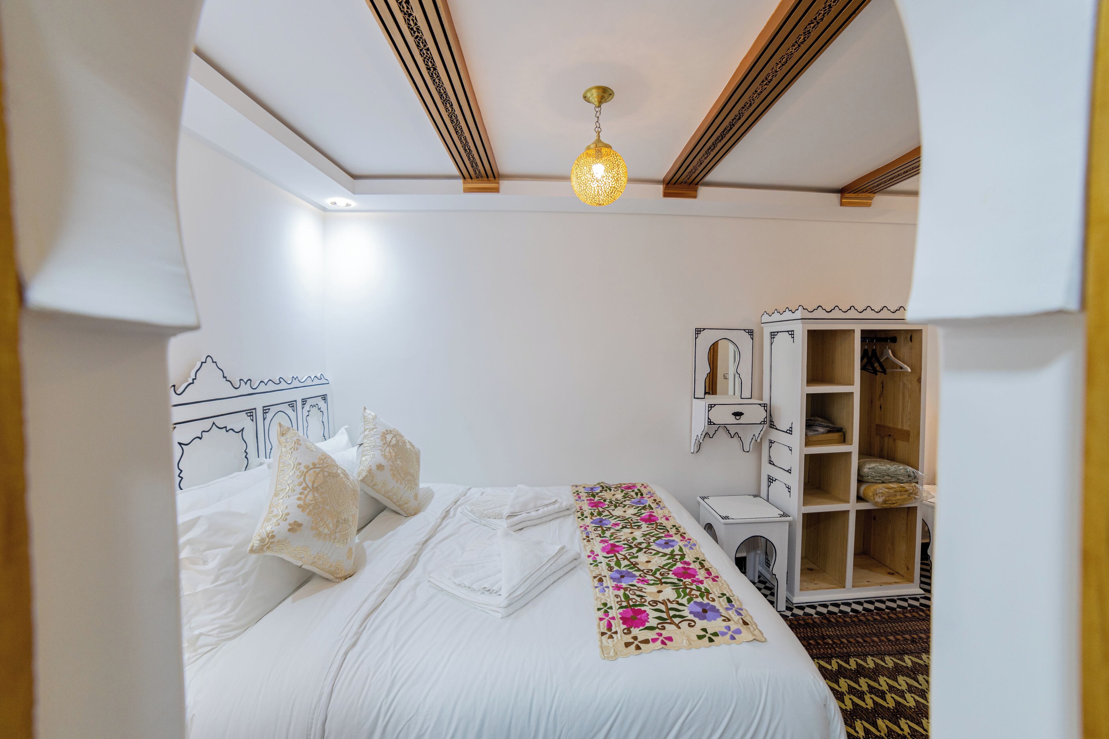 Una cama bien hecha con sábanas blancas y un corredor floral, un armario de madera y una lámpara colgante.