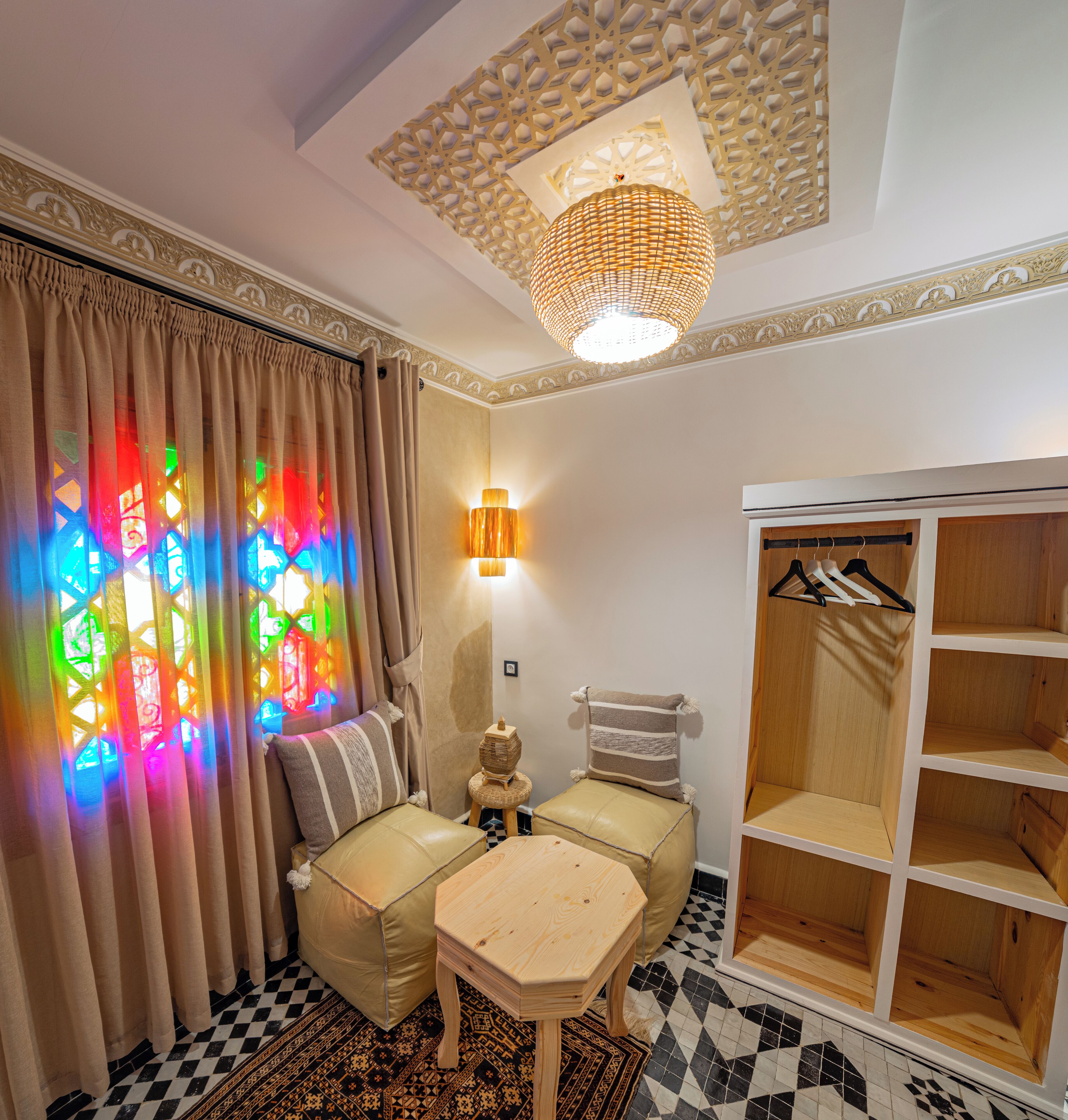 A room with a checkered floor, a wooden table, a sofa with cushions, a hanging light fixture, and a wardrobe with shelves.