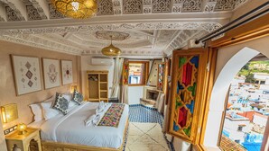 Ein Zimmer mit einem Bett, einem kleinen Tisch, einem dekorativen Teppich und Blick ins Freie.