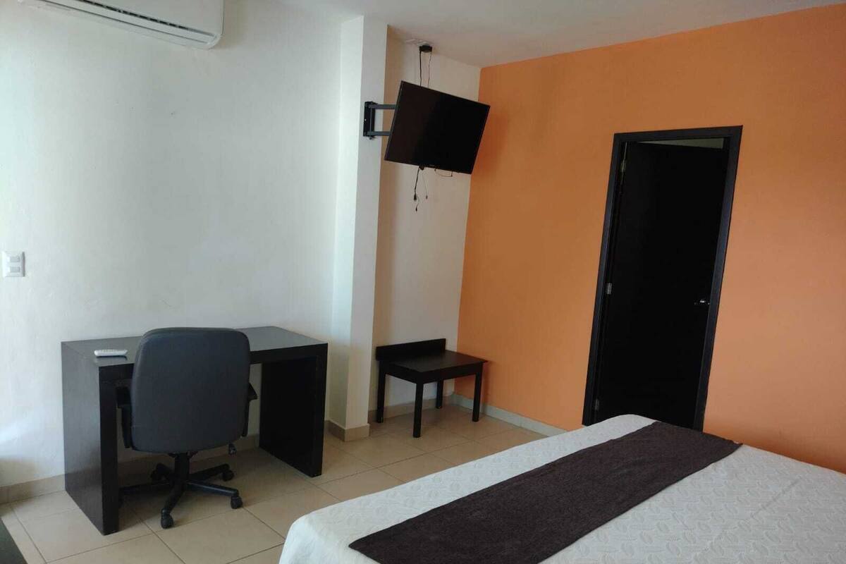 A hotel room with a bed, a desk, a chair, a TV mounted on the wall, and a door.