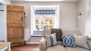 Ein gemütliches Wohnzimmer mit einem beige farbenen Sofa, gemusterten Kissen, einer Holztür und einem Fenster mit Blick auf Pflanzen und einen Zaun.