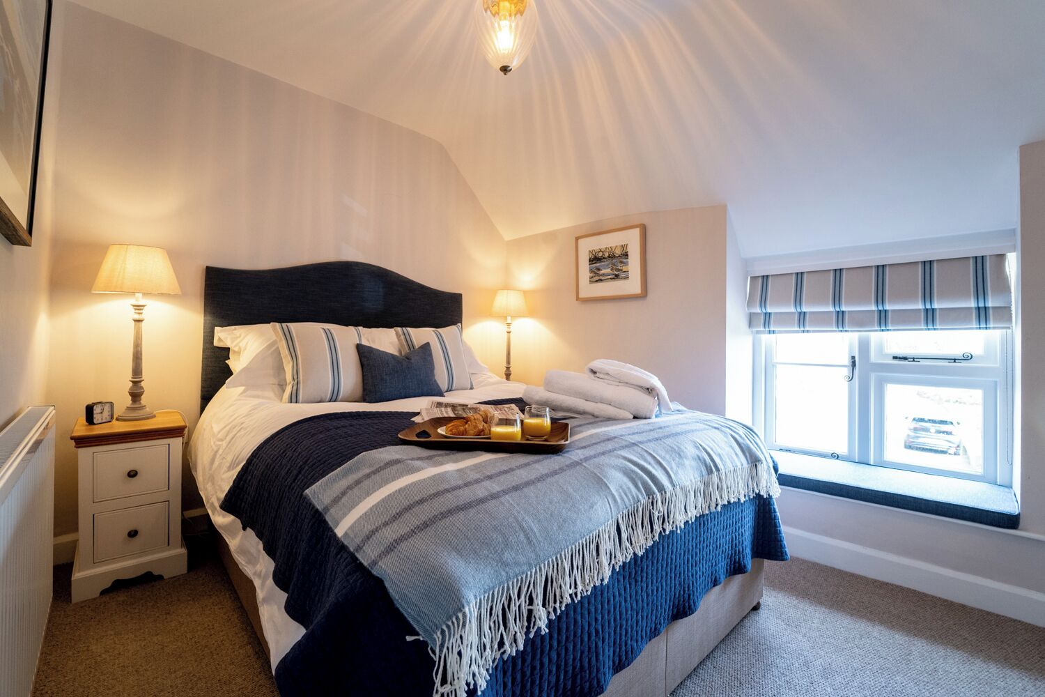 A neatly made bed with a tray of breakfast, two bedside lamps, and a window with blinds.