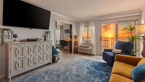 Un salon doté d’un téléviseur à écran plat, d’un meuble décoratif blanc, d’un tapis à motifs bleus, d’un canapé bleu et offrant une vue sur un coucher de soleil à travers une porte-fenêtre donnant sur un balcon.
