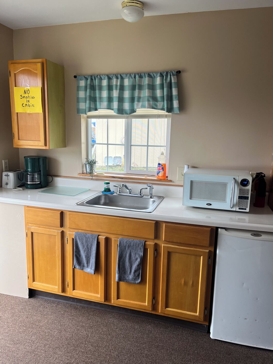 A compact kitchen with a sink, a microwave, and a refrigerator. There's a window with a checkered curtain, and a 'No Smoking' sign on the wall.