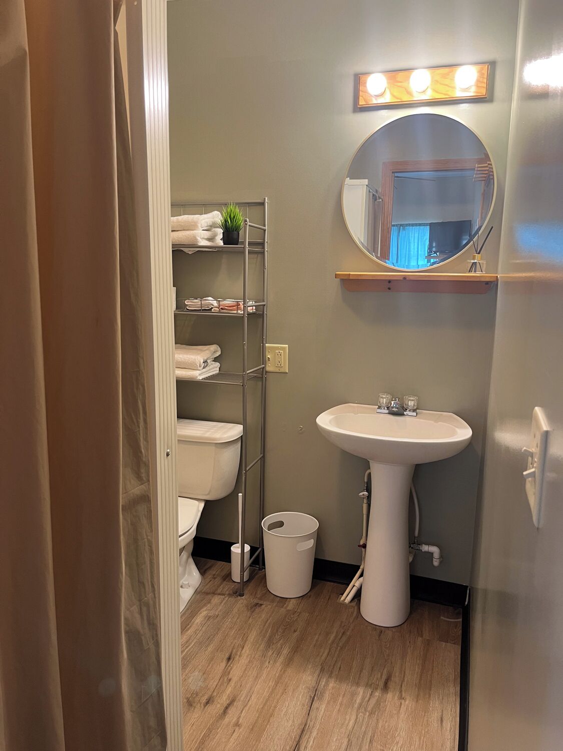 A compact bathroom with a white sink, a mirror with lights, a shelf with towels, and a small trash bin.