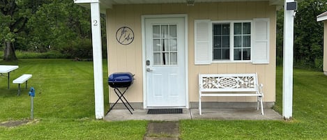 A small, single-story building with a beige exterior and white trim. It features a white door, a small porch with a bench, and a sign that reads "The Choo."