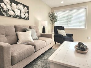 Ein Wohnzimmer mit einem beigen Sofa, einem schwarzen Sessel, einem weißen Couchtisch und einem Bild von weißen Blumen an der Wand.