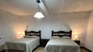 Une chambre avec deux lits, chacun doté d’une tête de lit en bois et d’une couvre-lit matelassé. On y trouve des tables de chevet avec des lampes, et la chambre a un plafond en pente avec un seul point lumineux.