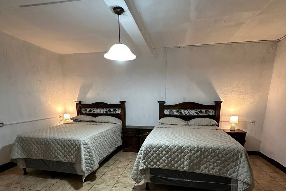 Una habitación con dos camas, cada una con cabecero de madera y colcha acolchada. Hay mesitas de noche con lámparas, y el techo inclinado tiene una única lámpara.