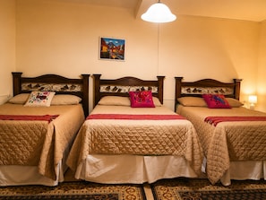 Une chambre d’hôtel avec trois lits, chacun doté d’une tête de lit en bois, de draps beiges et d’oreillers décoratifs. Un tableau est accroché au mur, au-dessus des lits.
