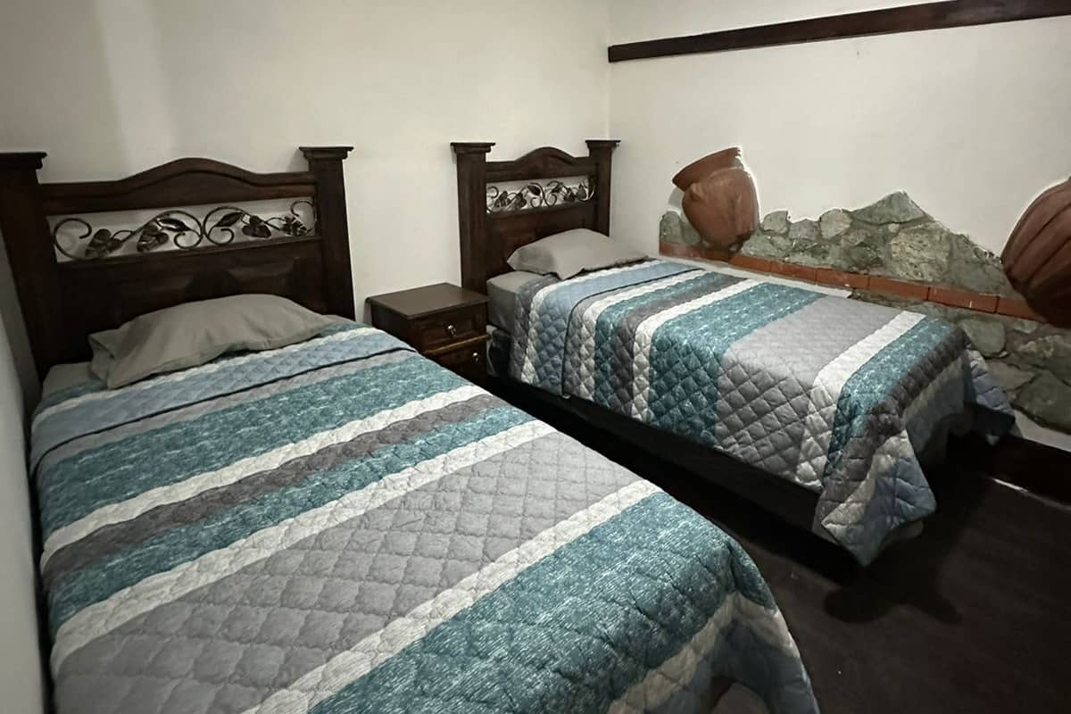 Un dormitorio con dos camas de madera, una mesita de noche, un cabecero decorativo y una pared de piedra.