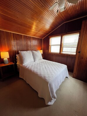 Une chambre avec un plafond en bois, un grand lit avec une literie blanche, deux lampes de chevet et une fenêtre avec des stores.