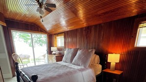 Ein Schlafzimmer mit Holzbalkendecke, einem Bett mit weißen Leinen, einem Nachttisch mit Lampe und einem Stuhl am Fenster.
