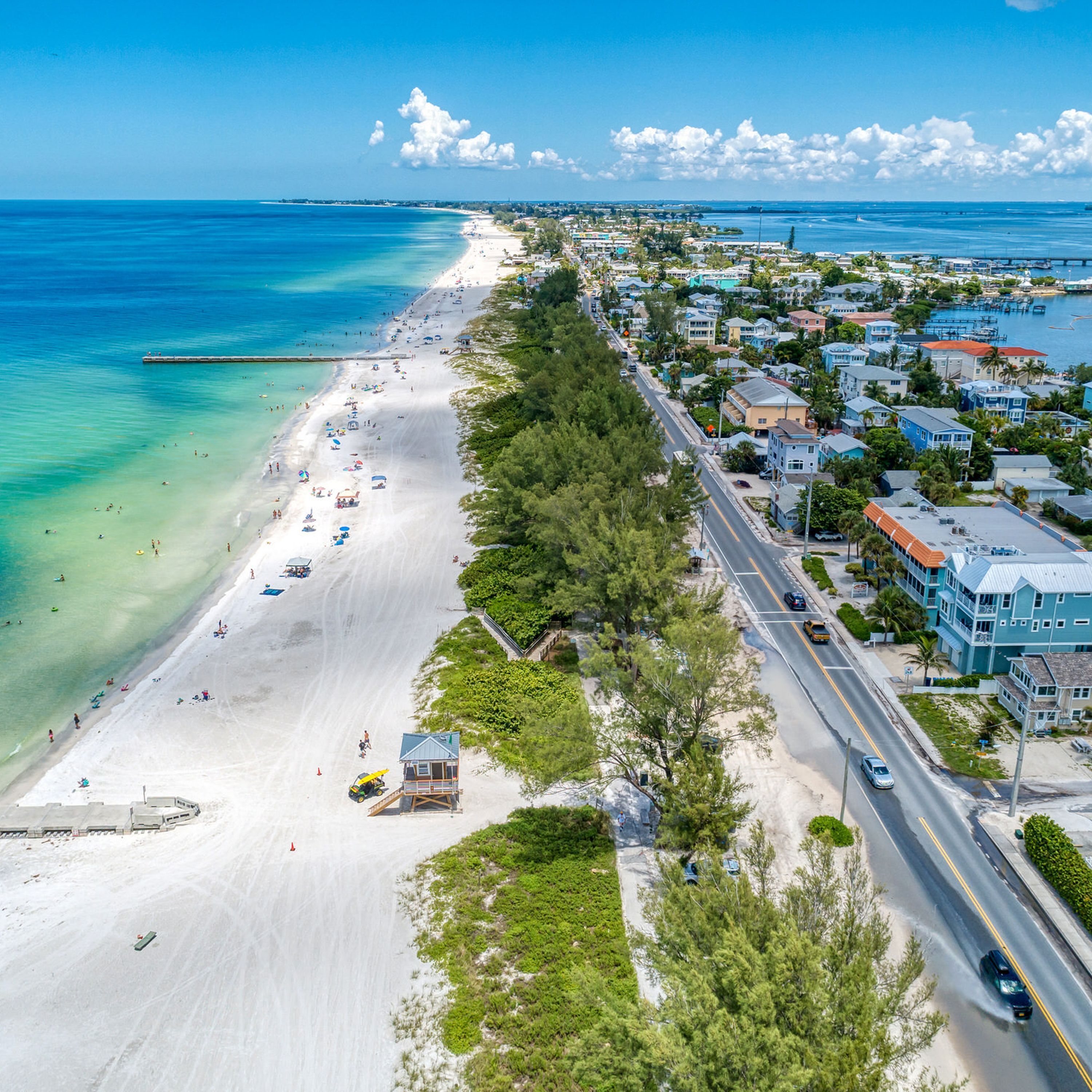 An aerial view of a coastal town with a long sandy beach, a road parallel to the shore, and residential buildings.