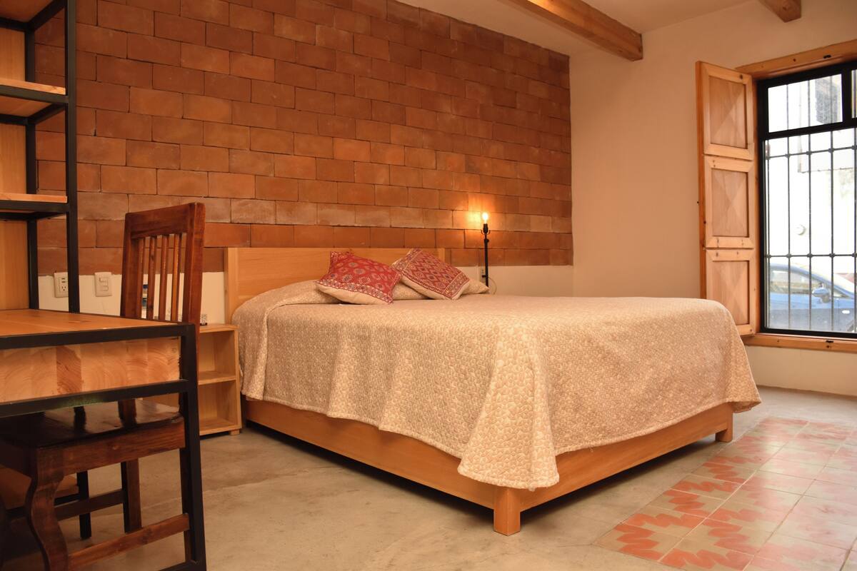 Ein Schlafzimmer mit Backsteinwand, einem hölzernen Bett, einem Nachttisch, einem Stuhl und einem Fenster mit Blick auf die Straße.