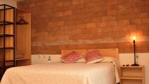 Een slaapkamer met een bakstenen muur, houten balken, een bed met bedkussens met een patroon, een nachtkastje met een lamp en een kledingkast.