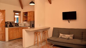 Een ruimtelijk ingerichte leefruimte met kitchenette, houten kasten, een grijze bank en een wandgemonteerde televisie.