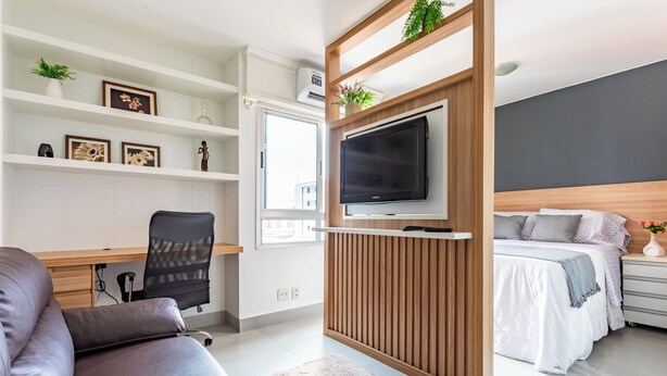 Un dormitorio moderno con una cama, un escritorio con silla, un televisor montado en un panel de madera y estantes con objetos decorativos.