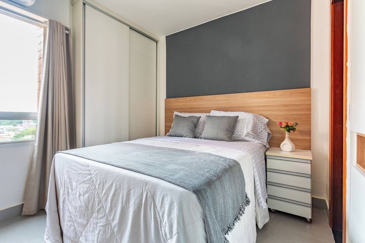 Ein ordentlich bezogenes Bett mit grauer Decke und weißer Bettwäsche, ein hölzernes Kopfteil, ein Nachttisch mit Blumen in einer Vase und Blick auf die Stadt durch ein Fenster.