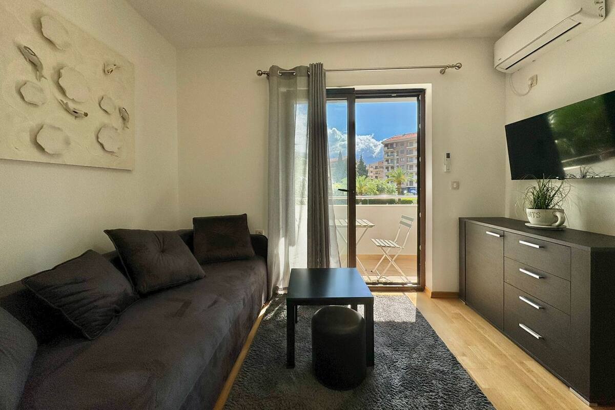 Un salón moderno con un sofá gris, una mesita y un armario. Hay un ventanal corredero que da a un balcón con vistas a edificios y árboles.