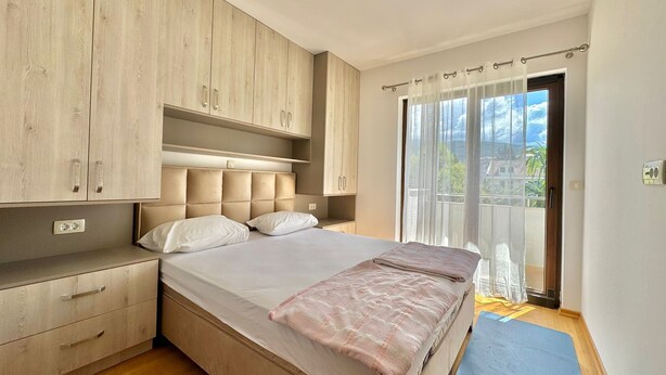 A bedroom with a bed, wooden cabinets, a window with curtains, and a view of the outside. - Central apartments Surina (Budva)