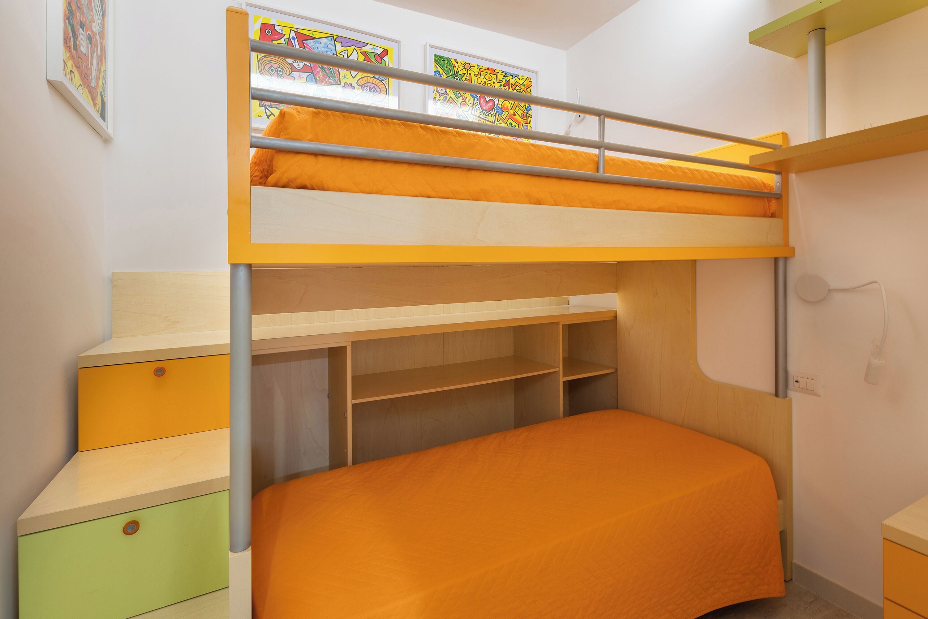 A bunk bed with a wooden frame and orange bedding, a small desk with a green drawer, and a wall-mounted shelf.