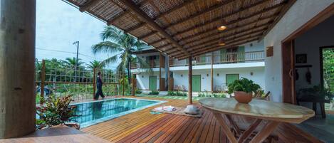 A wooden deck with a pool, a covered area with a thatched roof, and a view of a building with balconies and greenery.